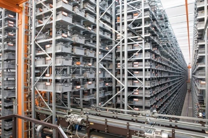 Automated high-bay warehouse with a miniload system, featuring tall storage racks filled with bins and trays, and conveyor systems for fast, high-density material handling and order picking.